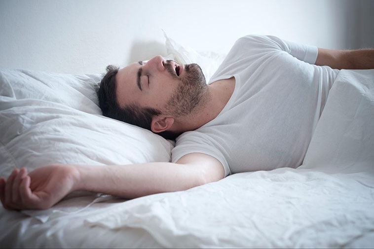 A young man sleeps sprawled on his bed, mouth open, with sleep apnea.